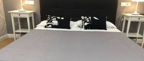 Una cama bien hecha con un colchón blanco y dos almohadas negras con diseños blancos, flanqueada por dos mesitas de noche con lámparas, en una habitación con paredes grises y suelo de madera.