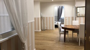 Habitación de hotel con piso de madera, una mesa de comedor con sillas, una cama con cubrecama estampado y un ventanal con cortinas.