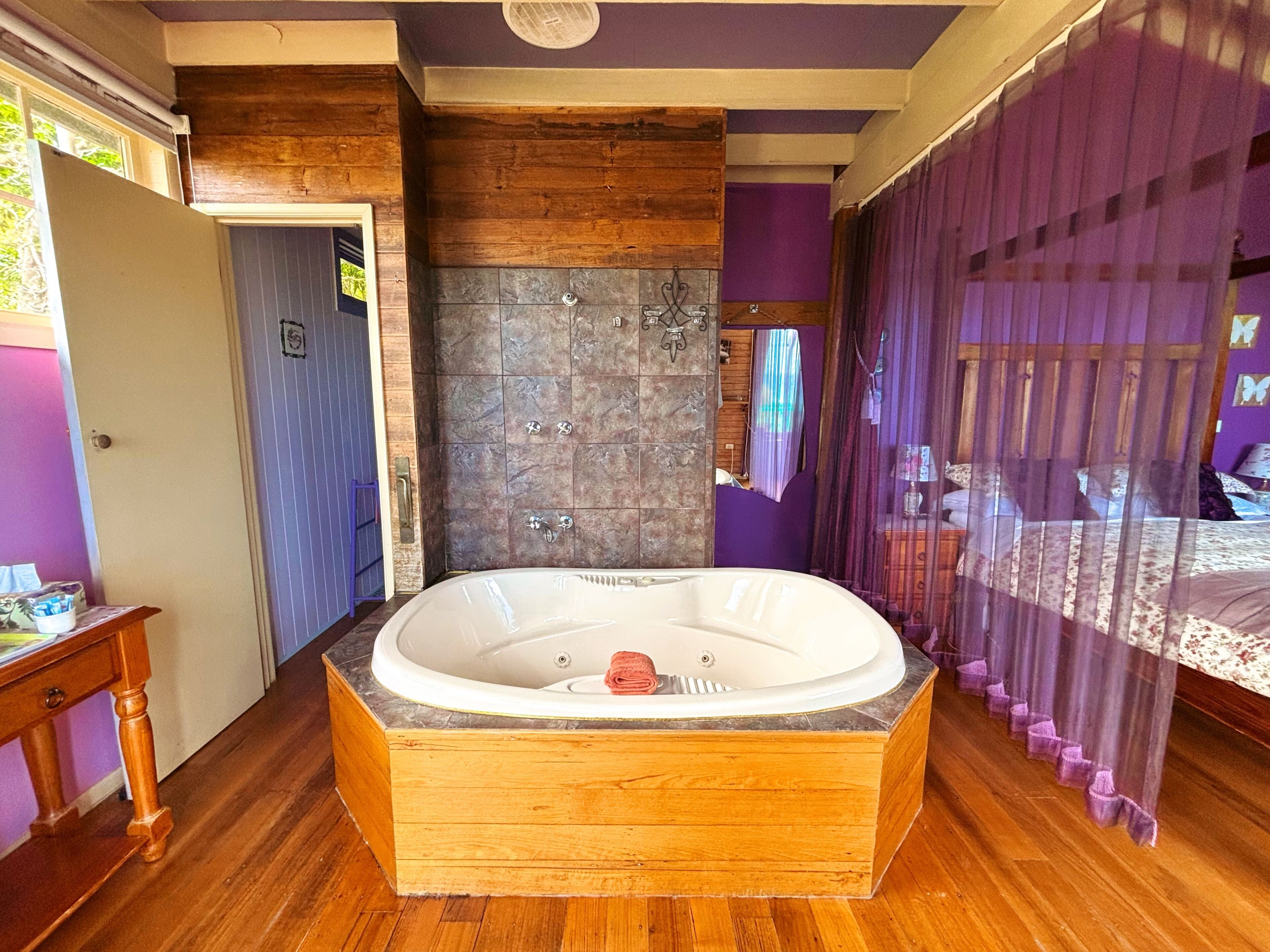 A bathroom with a wooden bathtub, a shower area, and a bed with purple curtains.