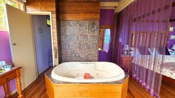 A bathroom with a wooden bathtub, a shower area, and a bed with purple curtains.