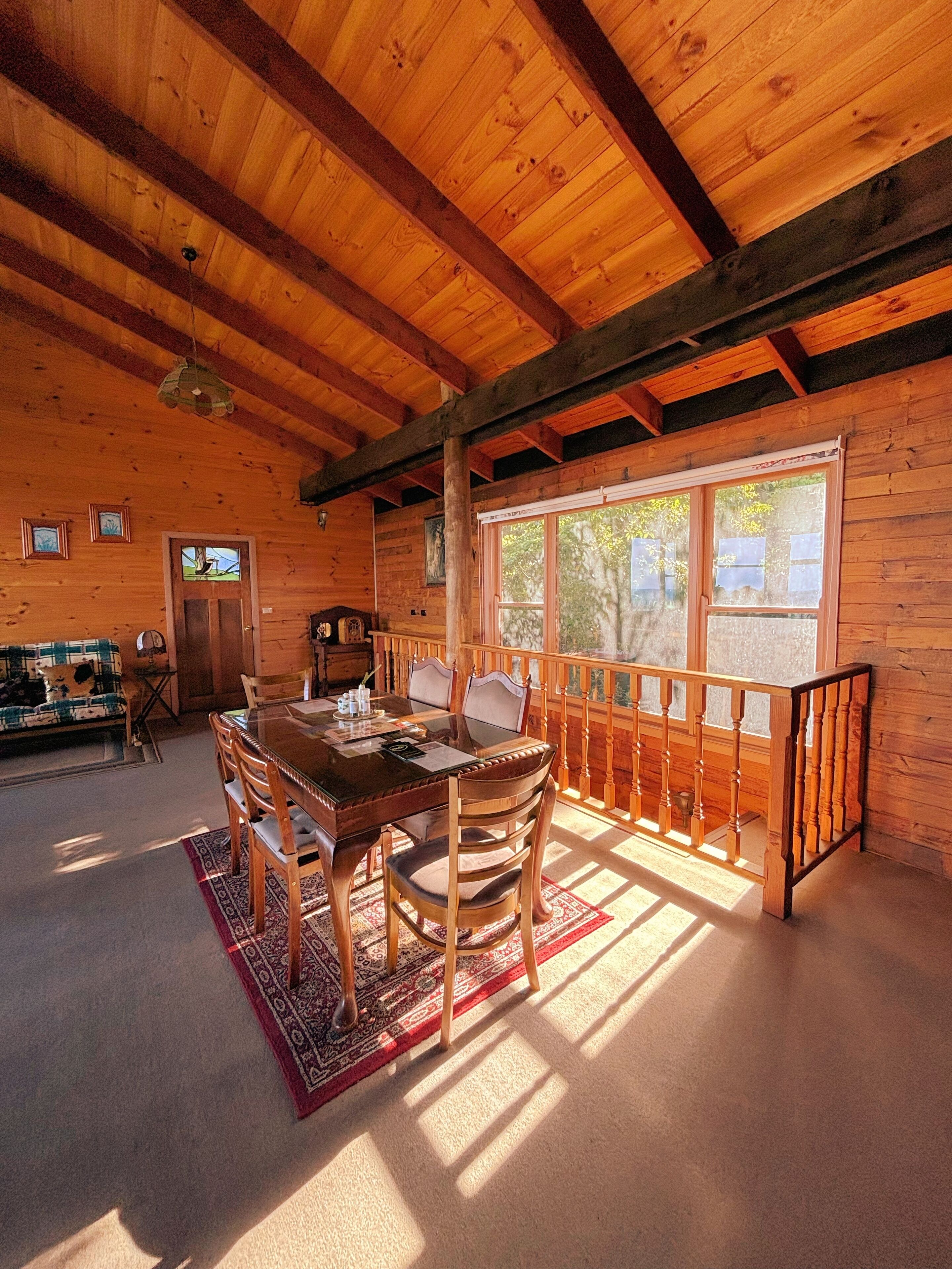 Interior de uma cabana de madeira com uma mesa de jantar, cadeiras, um sofá e uma janela com vista para árvores.