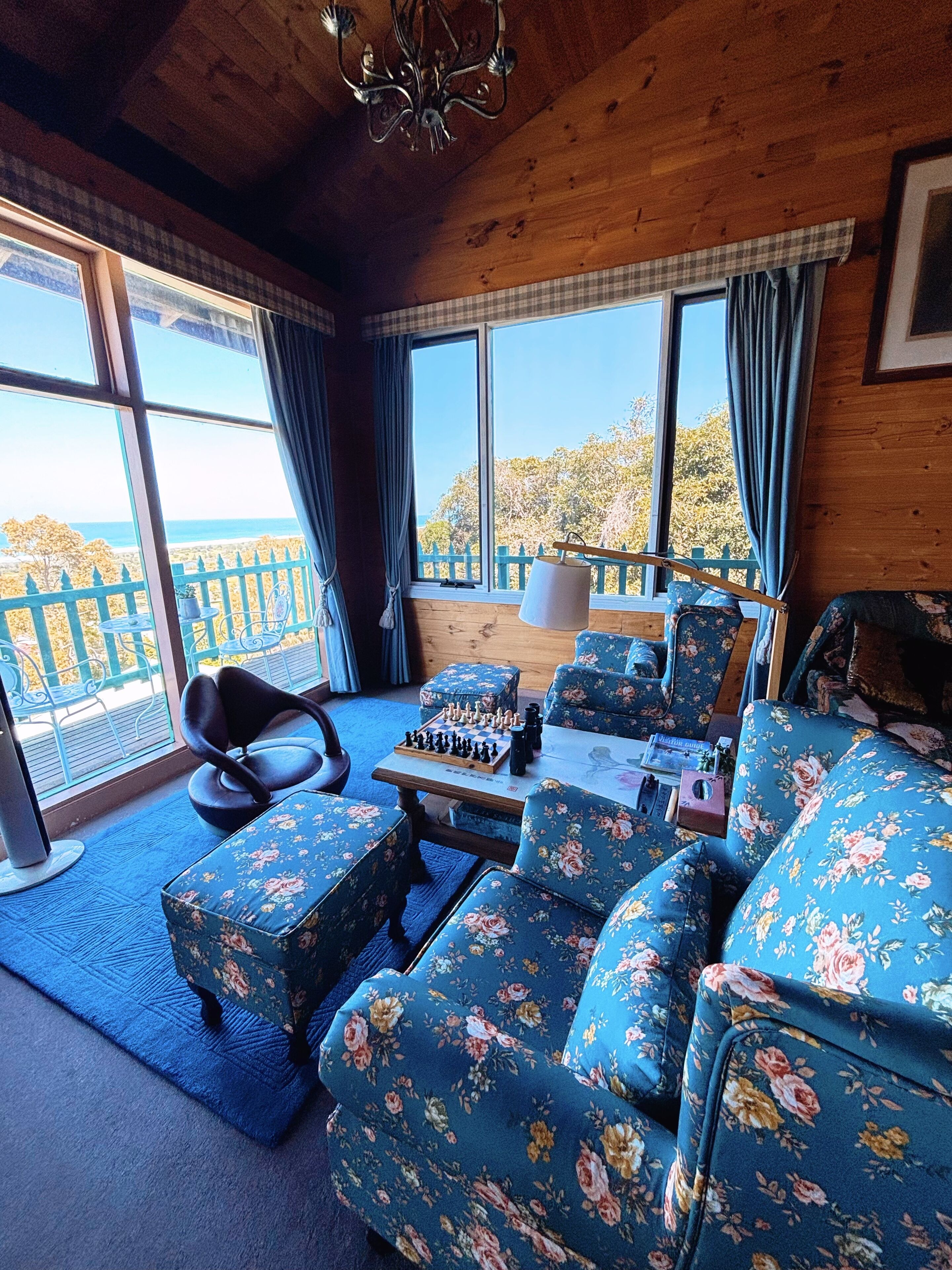 A cozy room with a wooden ceiling, large windows with curtains, a chessboard on a table, floral upholstered furniture, and a view of trees and a fence outside.