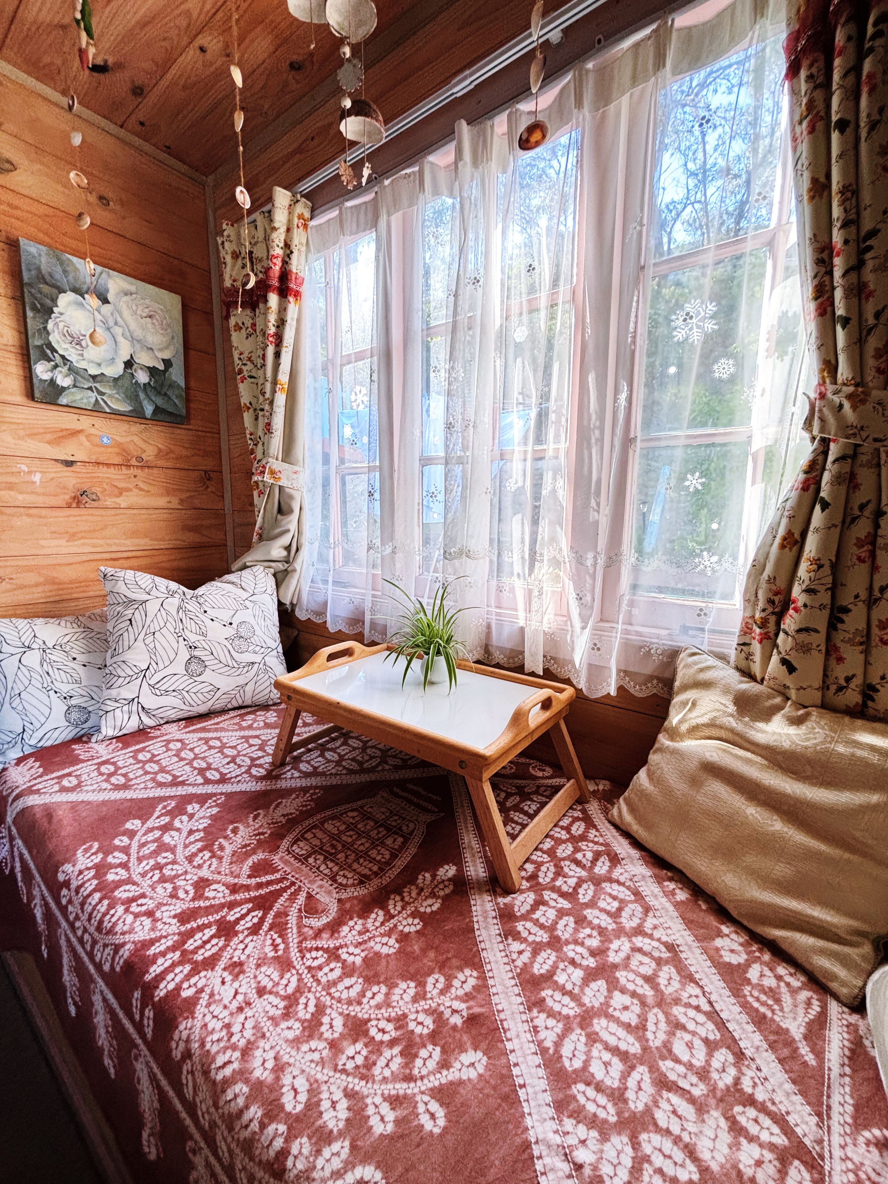 A cozy room with a wooden ceiling, a bed with patterned bedding, a small wooden table with a plant, and a window with floral curtains.