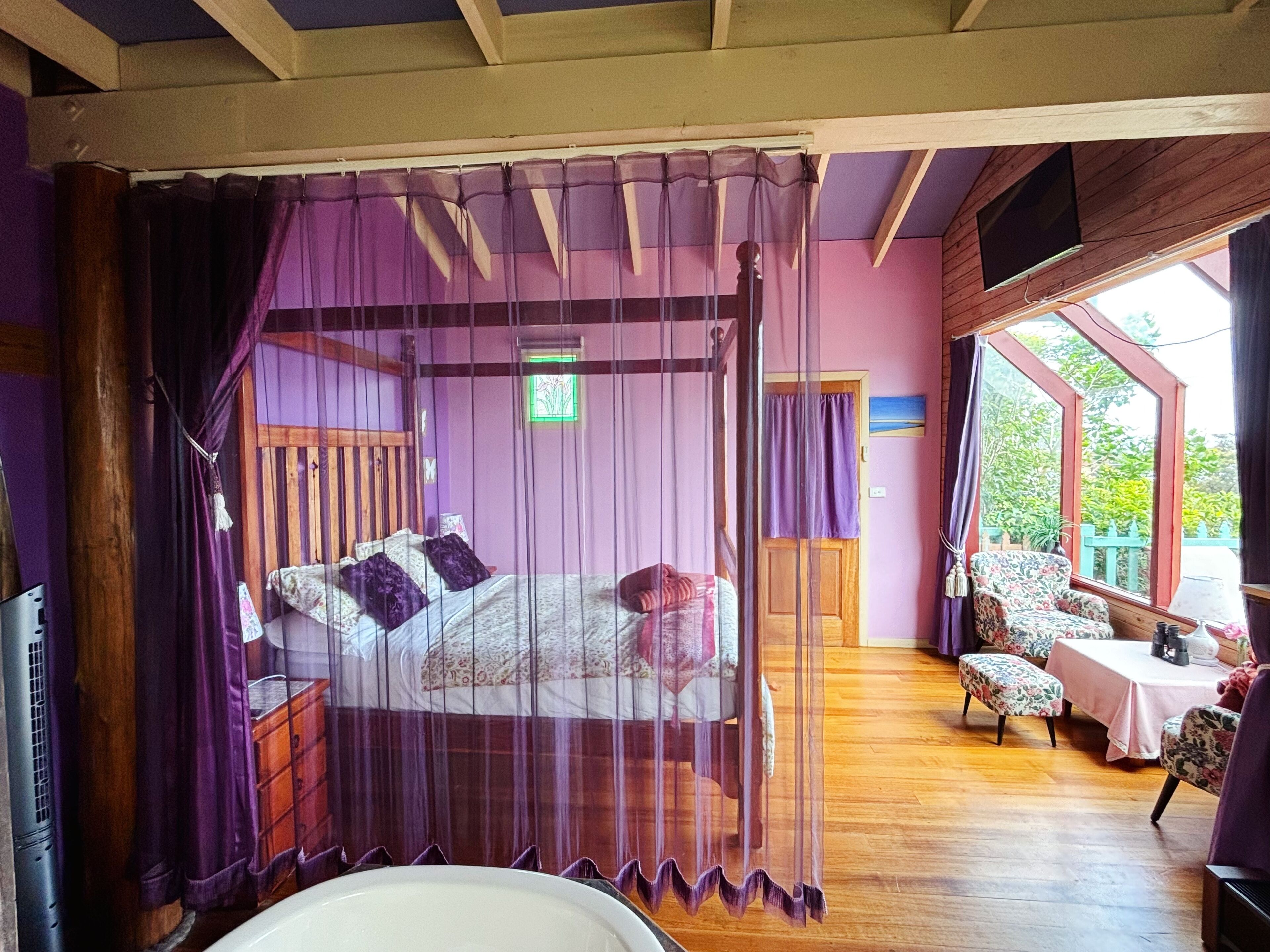 Ein Schlafzimmer mit Himmelsbett, Holzboden, einem kleinen Tisch mit Stühlen und Blick ins Freie.