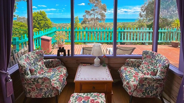 A room with a wooden floor, two floral-patterned chairs, a small table with a lamp, and a view of the ocean through large windows. - The Goat and Goose B&B (Lakes Entrance)