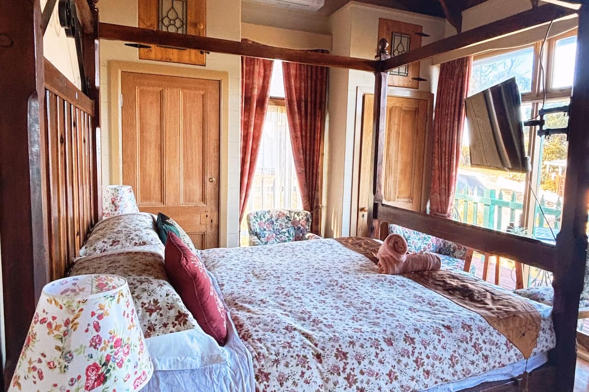 A wooden four-poster bed with floral bedding, a table lamp, and a view of the outdoors through a window.