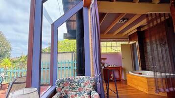 Quarto com uma janela grande, piso de madeira, uma poltrona floral, uma mesa com abajur e um banco de madeira.