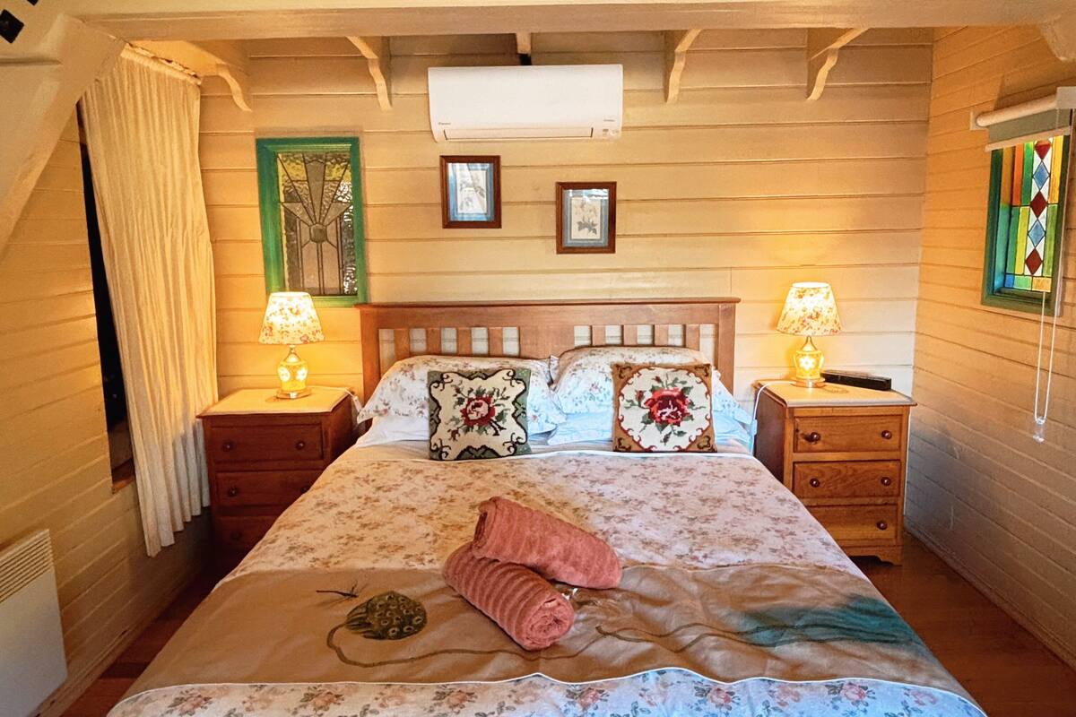 A bedroom with a bed, two bedside tables with lamps, a window with a stained glass design, and a ceiling fan.
