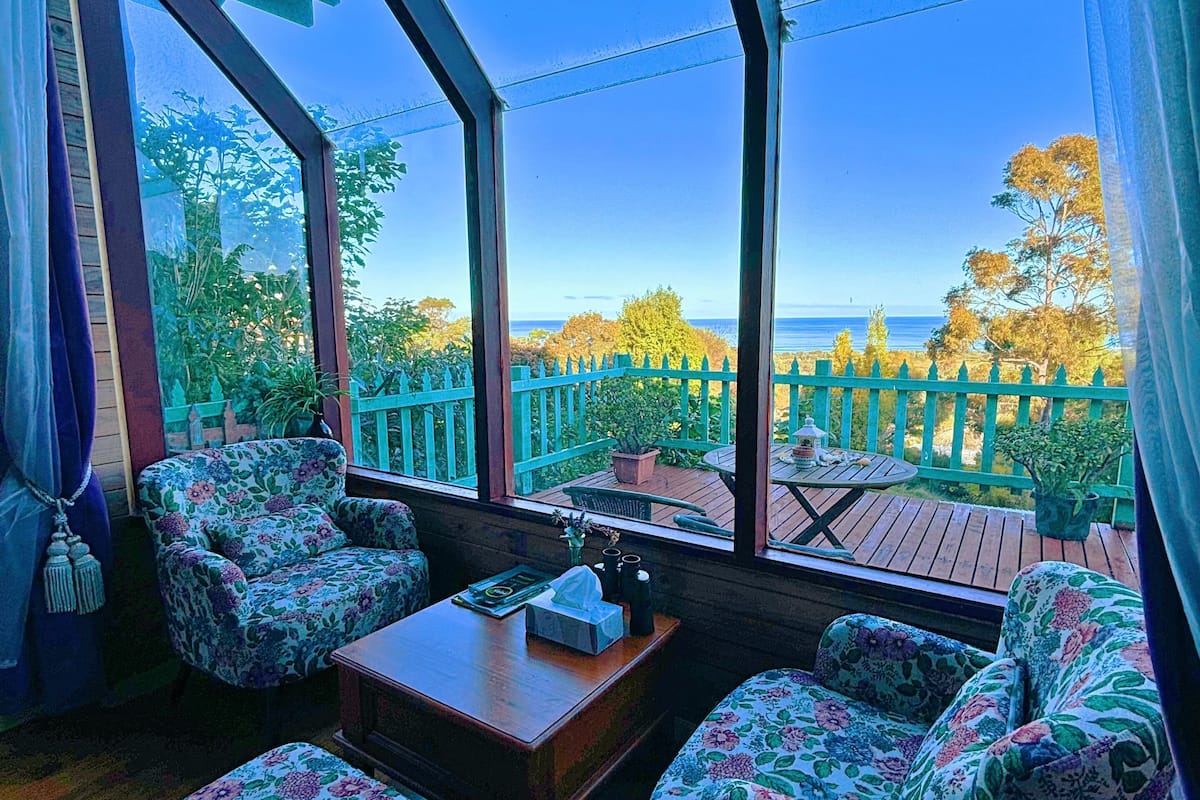 A room with a large window overlooking a deck with a green fence and trees. There are floral-patterned chairs and a small table with a tissue box.