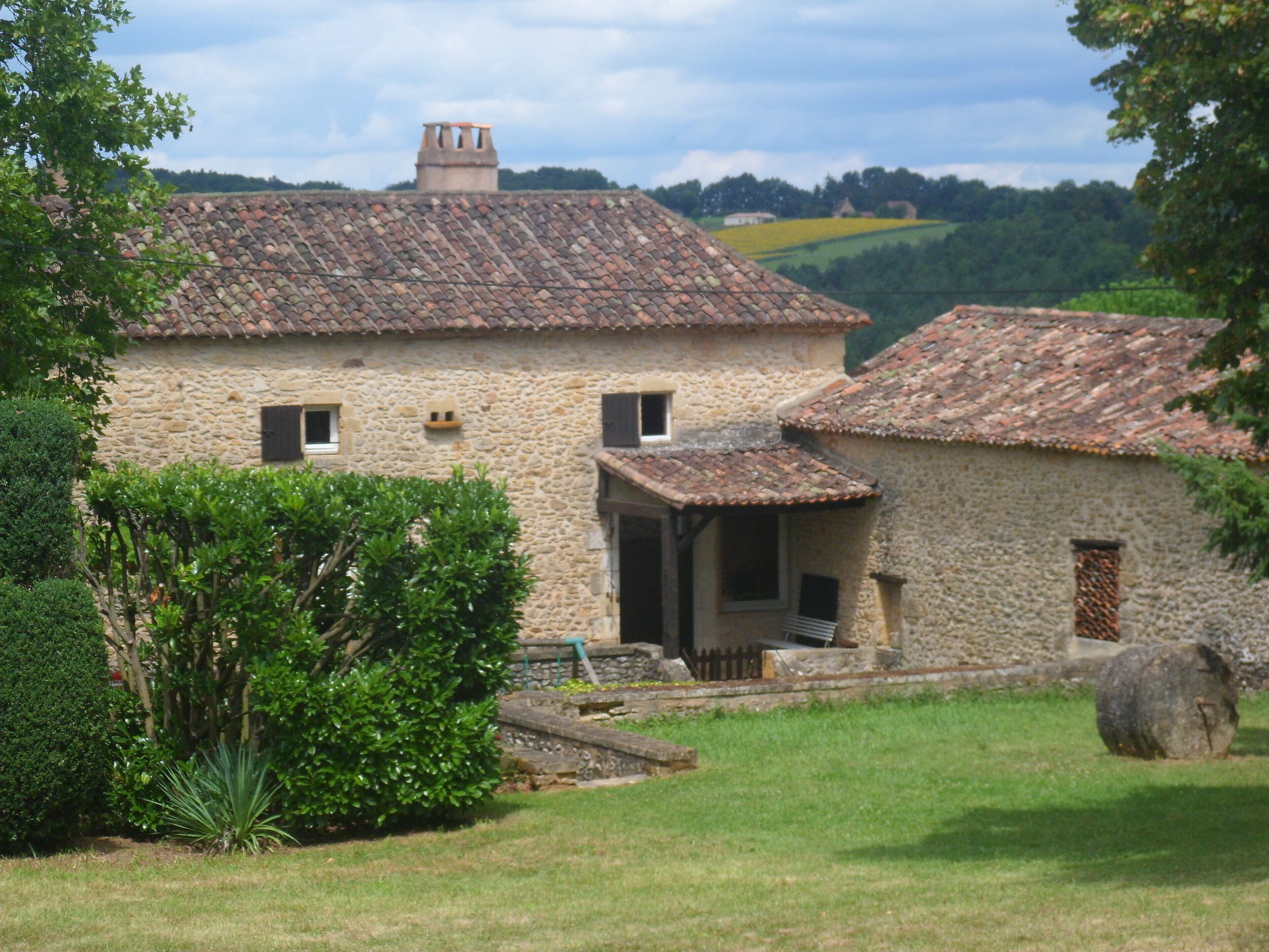 A stone building with a tiled roof, surrounded by greenery and a grassy area.