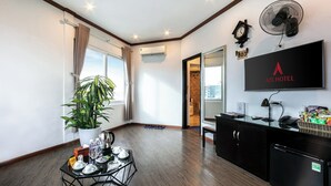 A hotel room with a wooden floor, a large potted plant, a glass table with plates and glasses, a black desk with a mirror, a TV, and a wall-mounted fan.