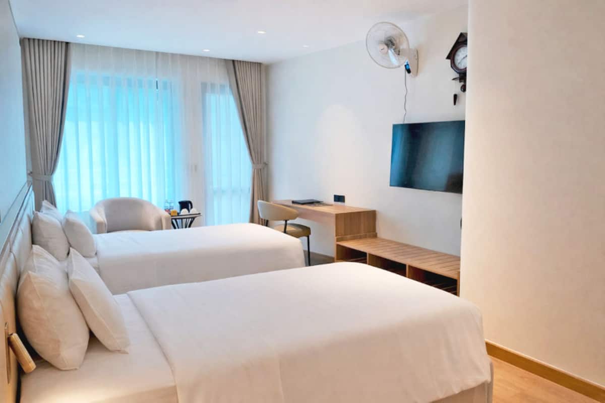 A hotel room with two beds, a desk, a chair, a TV, and a fan.