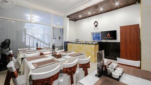 A hotel dining area with tables set for service, a reception desk, and a television screen displaying the hotel name.