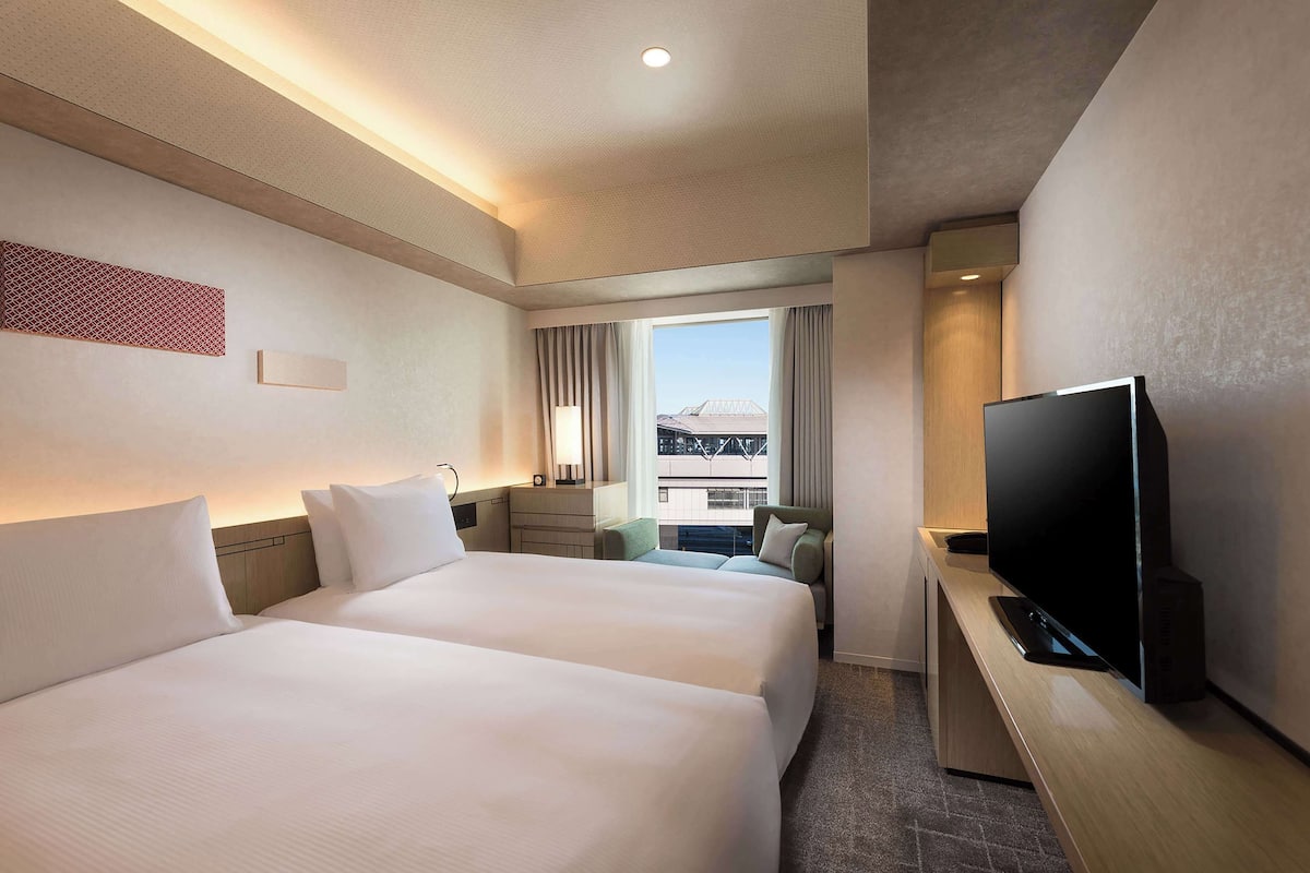 A hotel room with two beds, a TV, a desk, and a window with curtains.