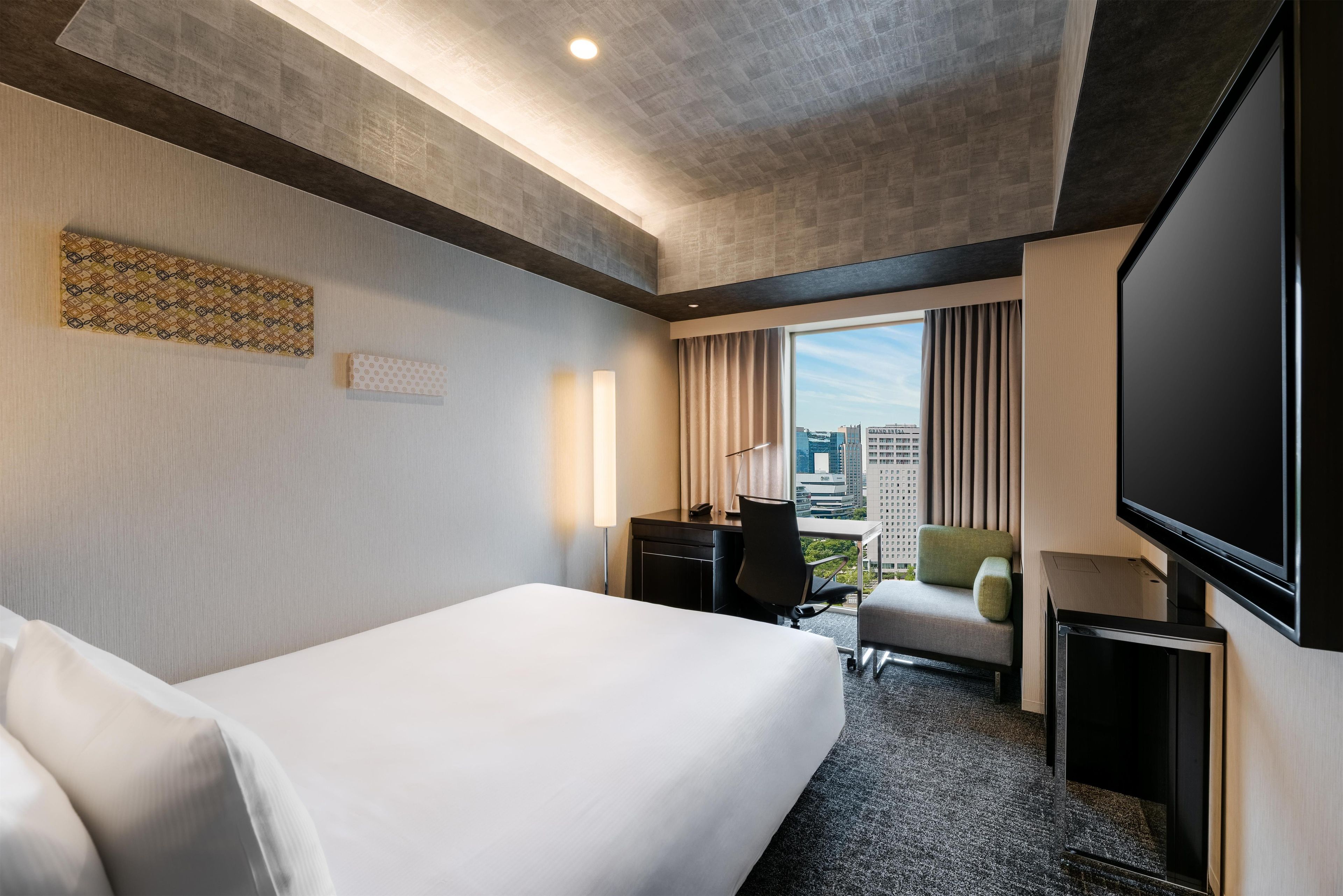 A hotel room with a large bed, a desk, a chair, a TV, and a window with a city view.