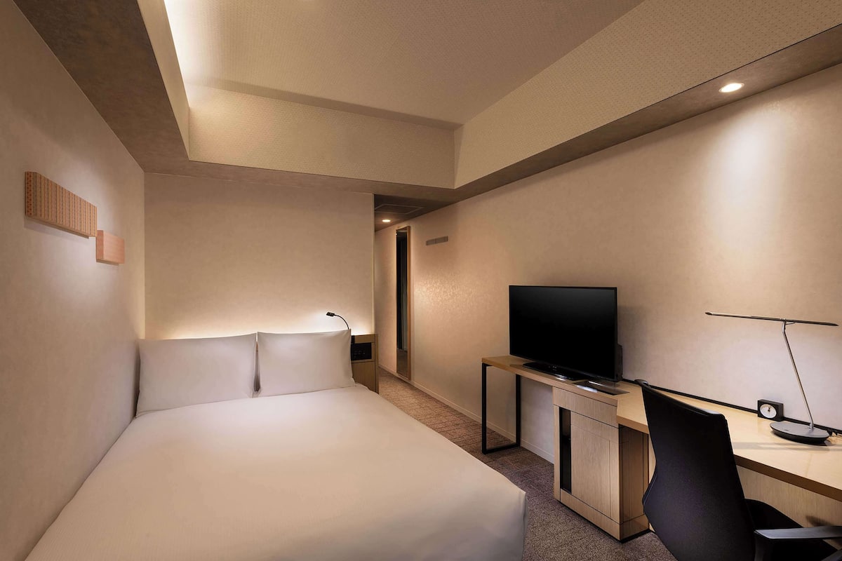 A hotel room with a bed, a desk with a television, and a chair.