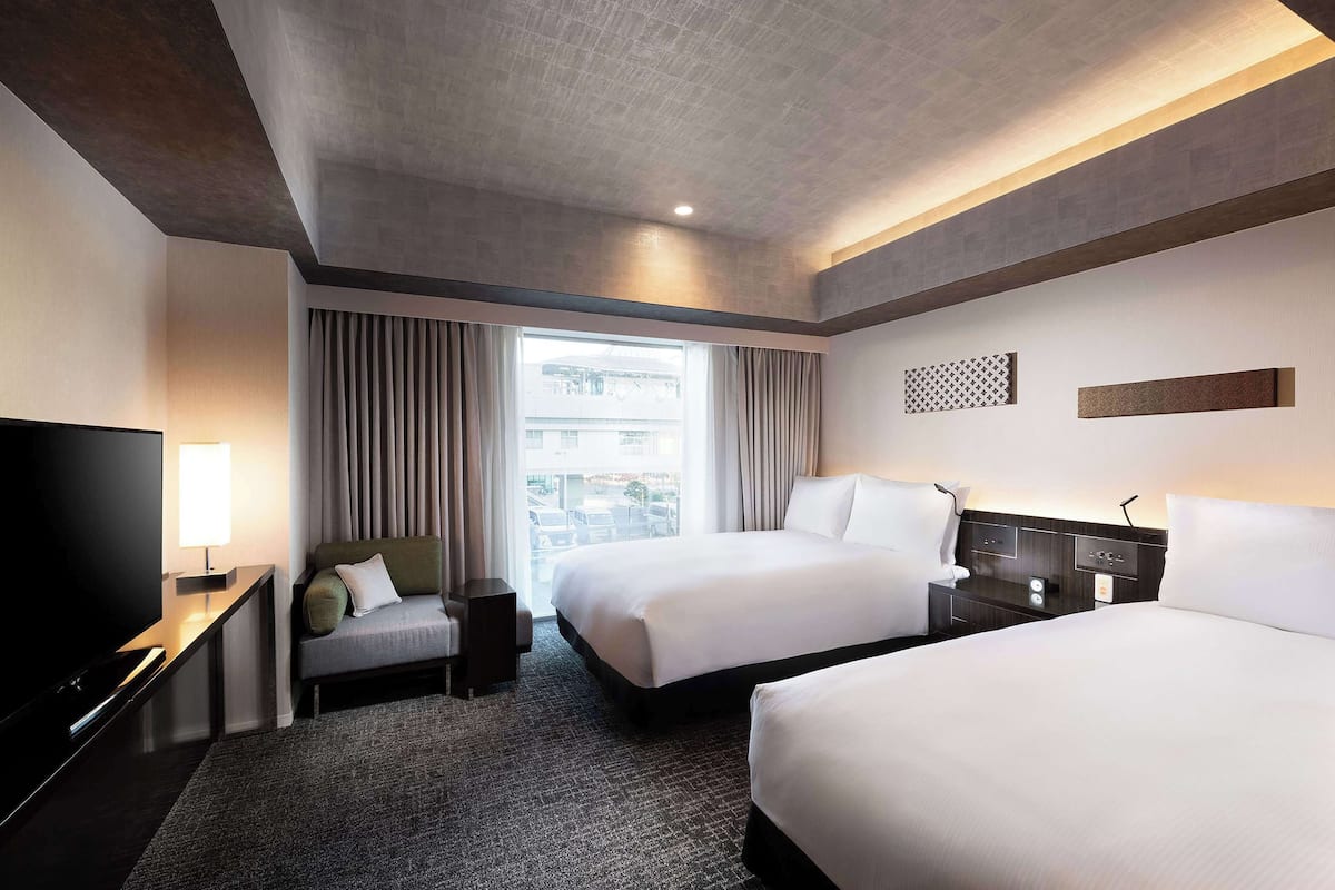 A hotel room with two beds, a TV, a desk, and a window with curtains.