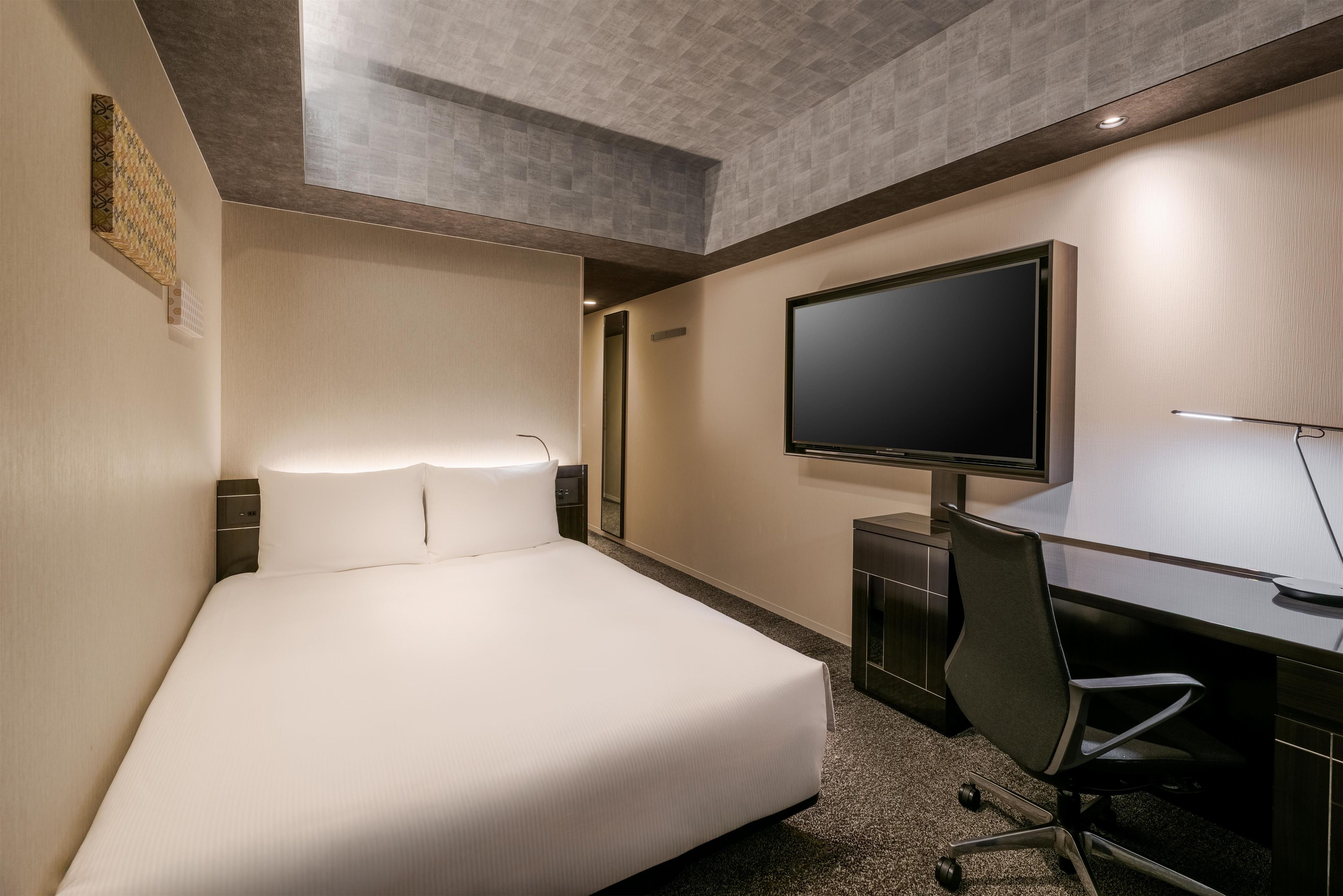 A hotel room with a bed, a desk with a chair, a flat-screen TV, and a mirror.