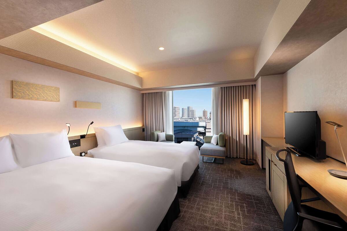 A hotel room with two beds, a desk with a TV, a chair, and a view of the cityscape.