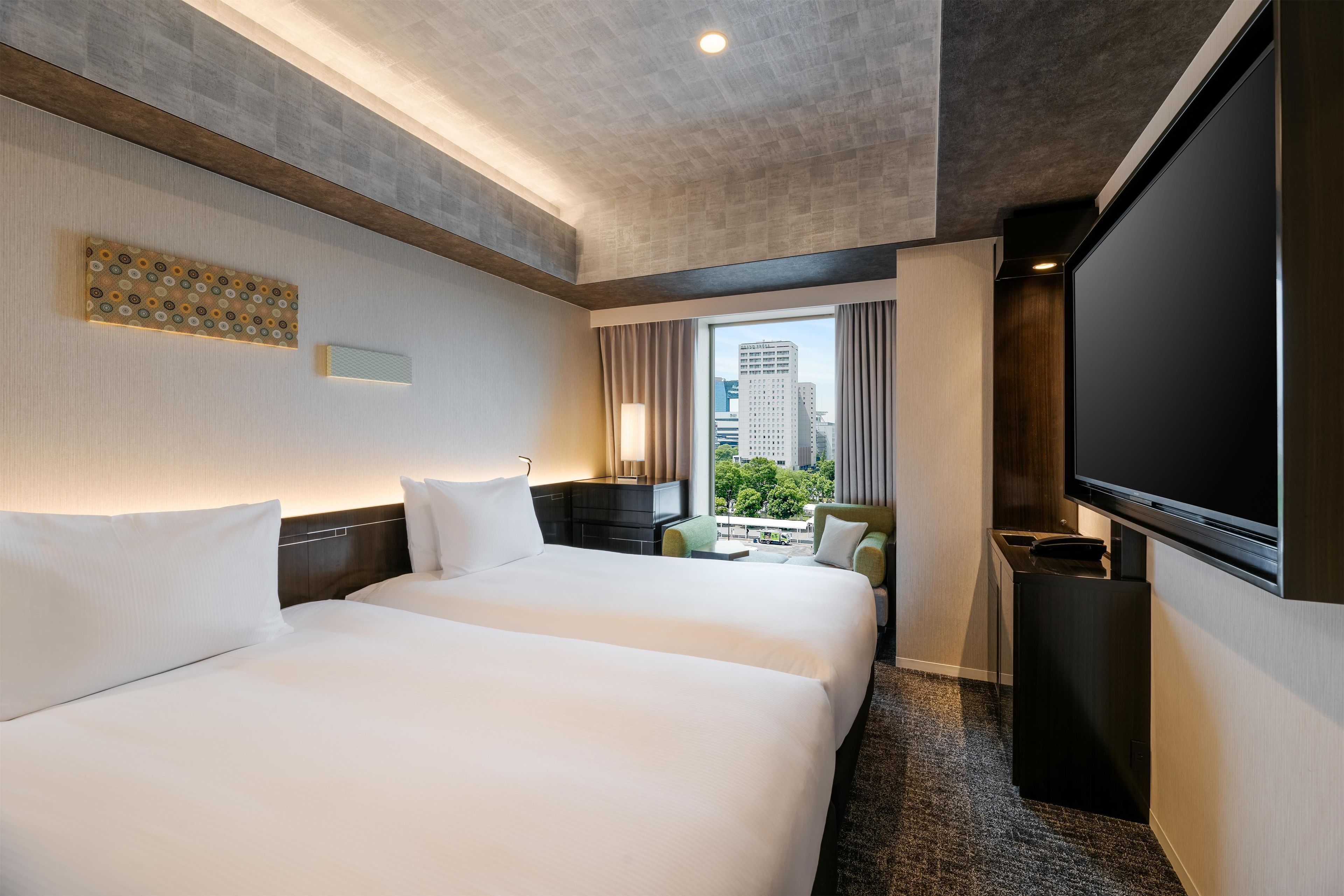 A hotel room with two beds, a TV, a desk, and a window with a city view.