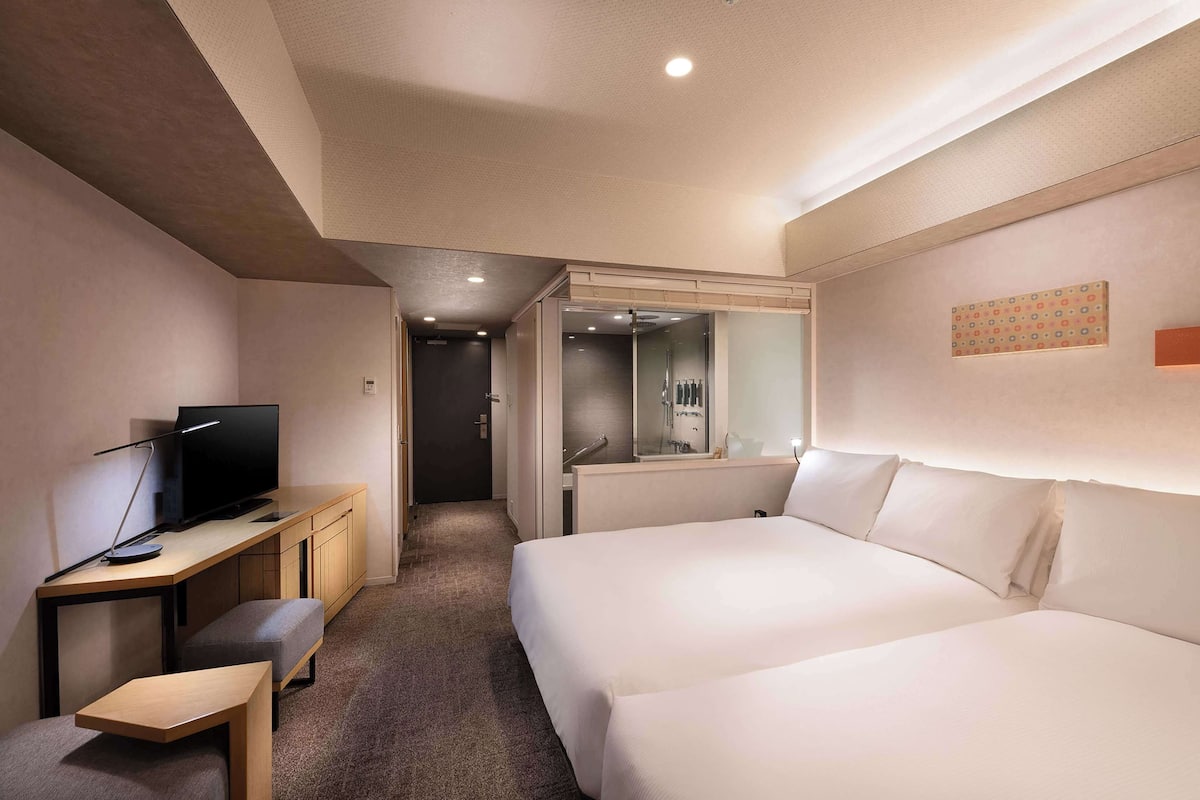 A hotel room with a bed, a desk with a computer, a chair, and a small table.