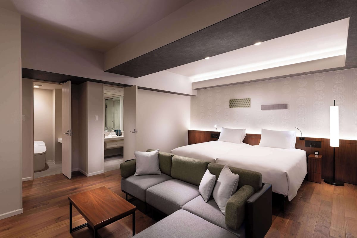 A modern hotel room with a large bed, a sofa, a small table, and a bathroom visible through an open door.