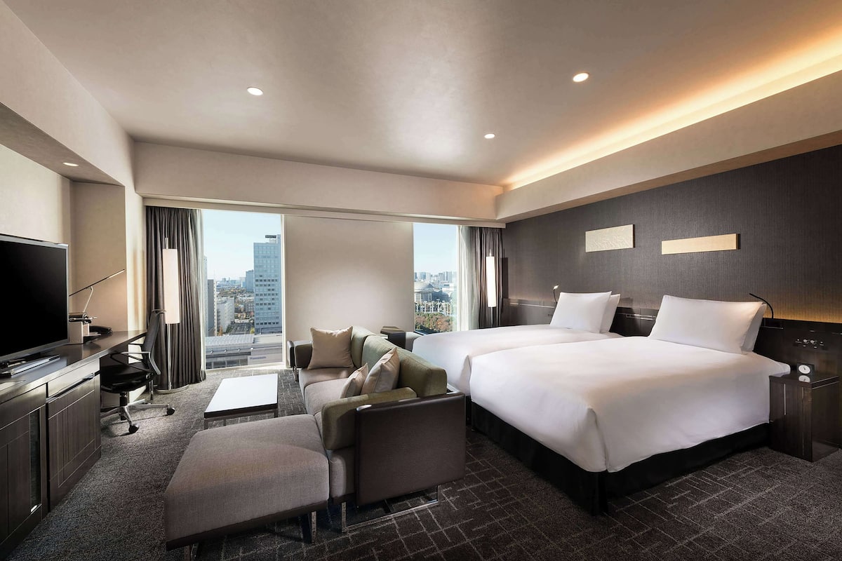 A hotel room with a large bed, a sofa, a desk with a TV, and a city view.