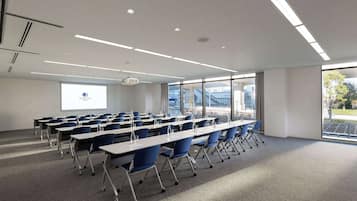 Une salle de conférence avec des rangées de tables et de chaises, un écran de projection et de grandes fenêtres donnant sur des bâtiments et des arbres.