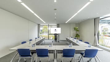 A conference room with a long table, blue chairs, a projector screen, and a potted plant.