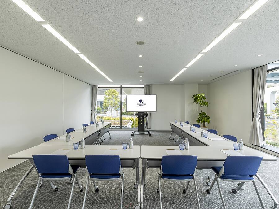 A conference room with a long table, blue chairs, a projector screen, and a potted plant.