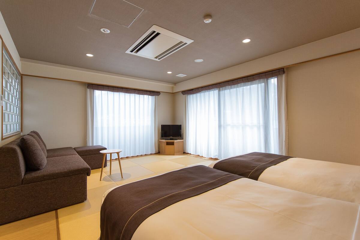 A modern hotel room with a sofa, two beds, a TV, and large windows.