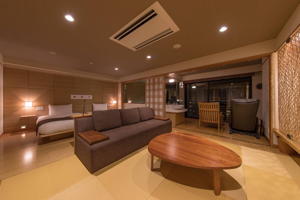 A modern hotel room with a sofa, a wooden coffee table, a bed, a small table with a lamp, a bathroom with a sink and toilet, and a sliding glass door leading to a balcony.