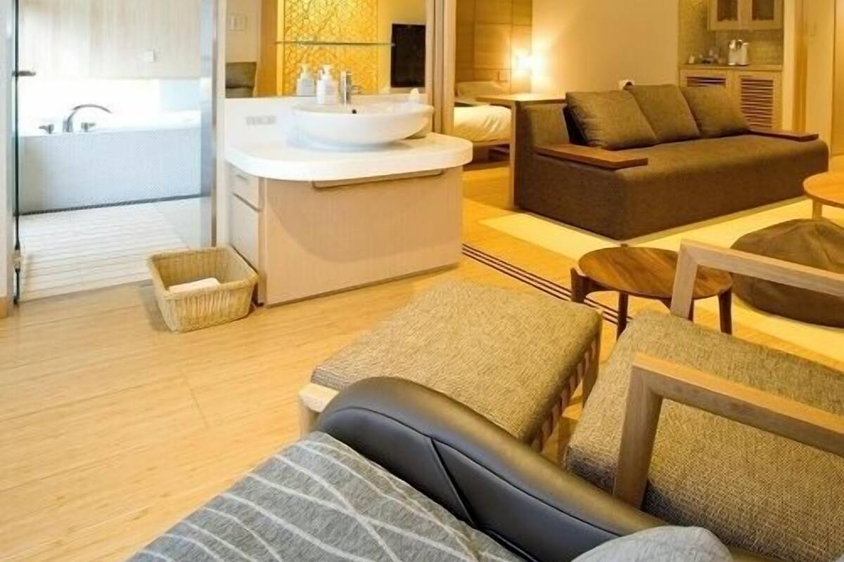 A modern hotel room with a sofa, a small table, a kitchen area, and a bathroom.
