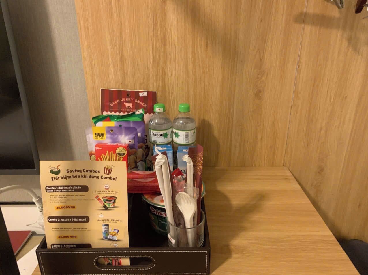 A wooden cabinet with a box of snacks, a bottle of Dasani water, and a glass with utensils.
