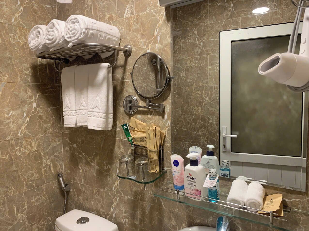 A bathroom with marble walls, a toilet, a glass shelf with toiletries, a towel rack, and a mirror.