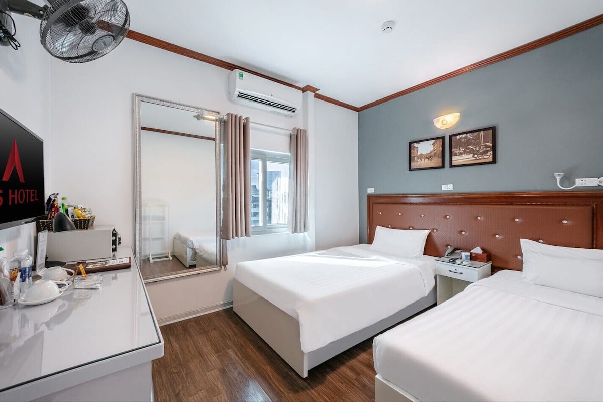 Ein Hotelzimmer mit zwei Betten, einem Schreibtisch mit Spiegel, einer Wand-Klimaanlage und einem Fenster mit Vorhängen.