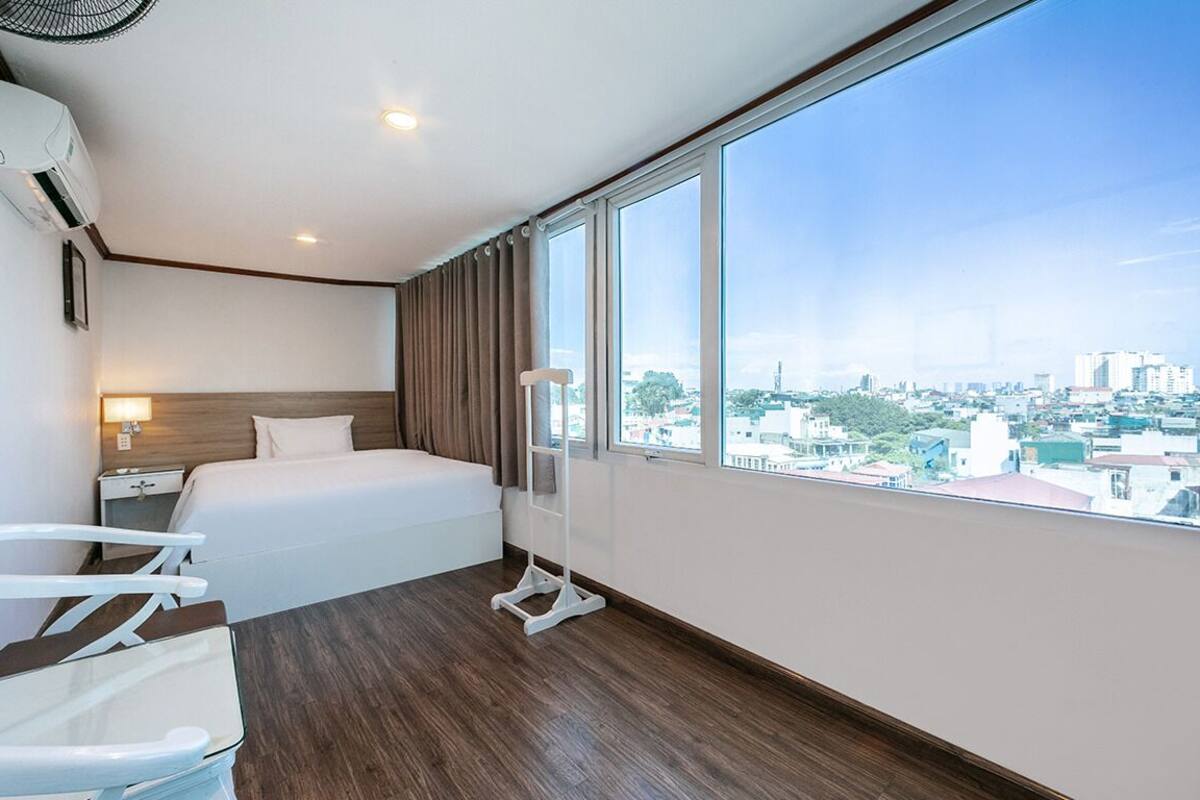 Ein Hotelzimmer mit einem großen Bett, einem Nachttisch, einem Sessel und Blick auf die Stadt durch große Fenster.