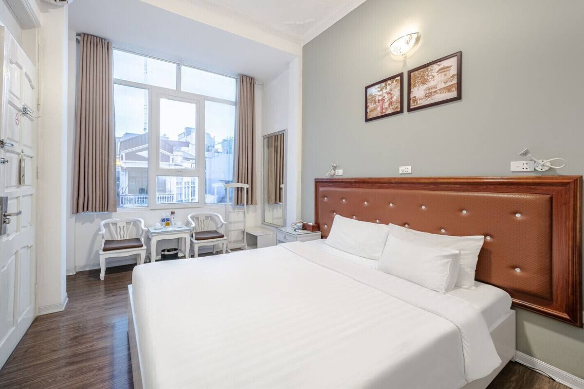 Ein Hotelzimmer mit einem großen Bett, einem Schreibtisch mit Stuhl, einem Fenster mit Vorhängen und zwei gerahmten Bildern an der Wand.