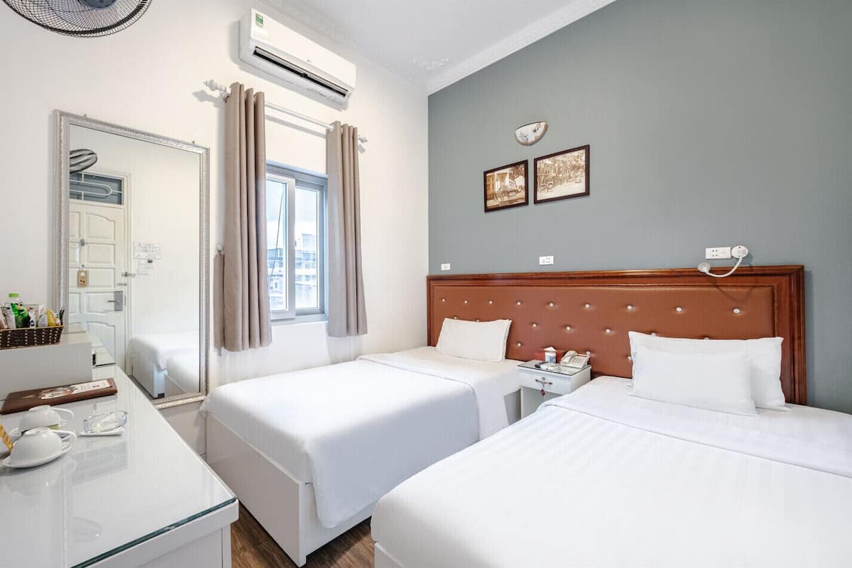Ein Hotelzimmer mit zwei Betten, einem Schreibtisch mit Spiegel, einer Klimaanlage und einem Fenster mit Vorhängen.