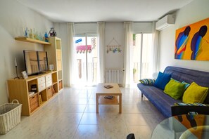 Sala de estar con sofá, una mesita de centro y un mueble para televisor. Se ve una ventana con cortinas, un radiador y una obra de arte colgada en la pared.