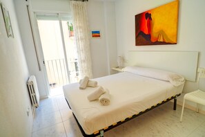 A neatly made bed with rolled towels, a white nightstand with a lamp, and a painting on the wall.