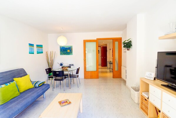 Una sala moderna con sofá, mesa de centro y mueble para la televisión. Hay un comedor con sillas y una mesa, y un pasillo que conduce a otra habitación.