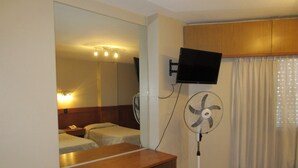A hotel room with a bed, a desk, a chair, a TV mounted on the wall, a standing fan, and a window with curtains. - Hotel Royal (Córdoba)