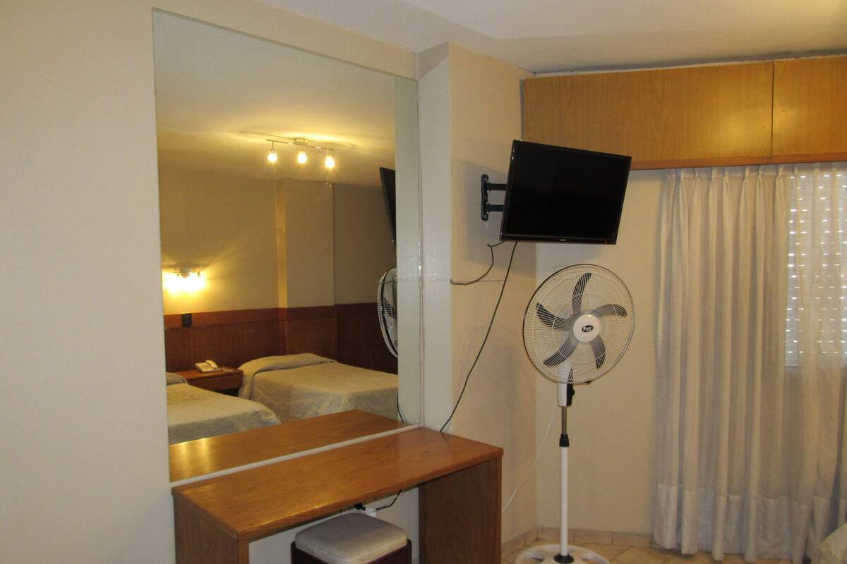 Habitación de hotel con cama, escritorio, silla, televisor montado en la pared, ventilador de pie y ventana con cortinas.