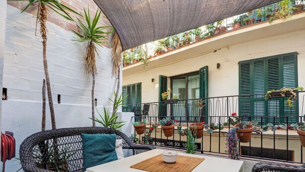 A patio with wicker furniture, a white table, and a small rug. There are potted plants and a balcony with green shutters. - Charming Patio by Hello Homes Sitges (Sitges)