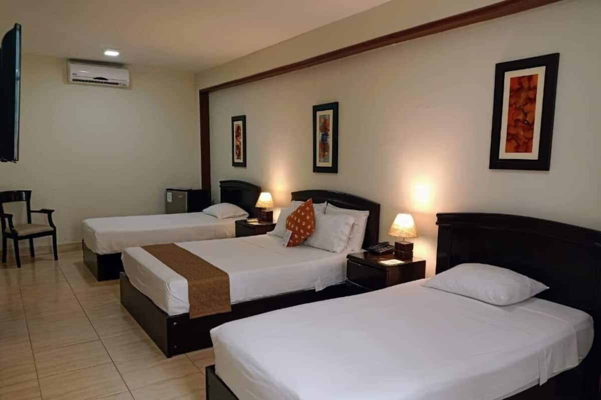 A hotel room with three single beds, a TV, an air conditioner, and framed artwork on the walls.