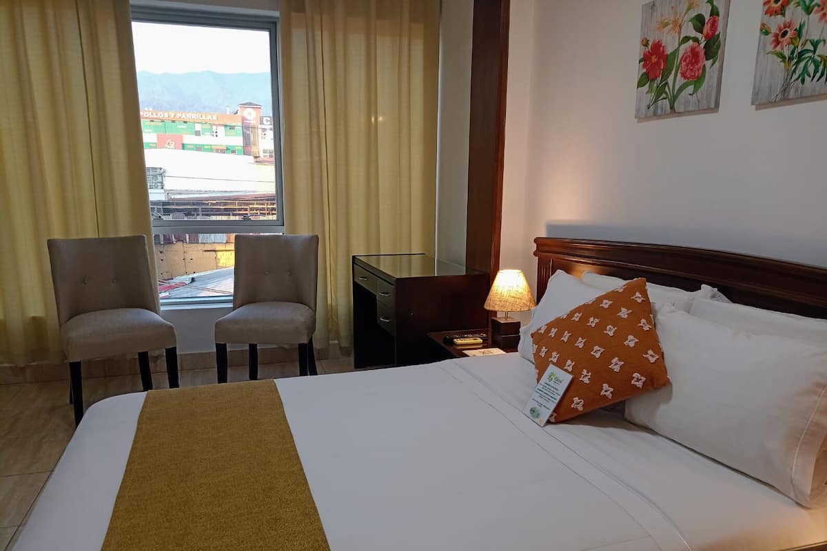 A hotel room with a bed, two chairs, a nightstand with a lamp, and a window with curtains.