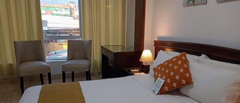 A hotel room with a bed, two chairs, a nightstand with a lamp, and a window with curtains.