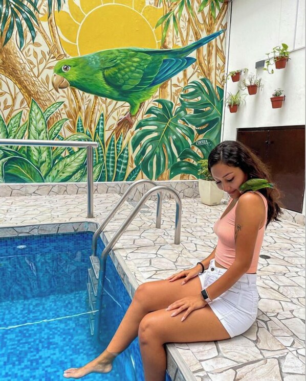 Una mujer sentada junto a una piscina, con un mural tropical de fondo.