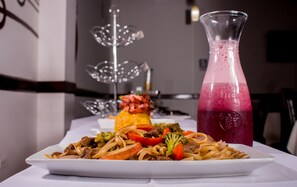 A plate of stir-fried noodles with vegetables and meat, a glass of pink beverage, and a decorative metal tree on a table.